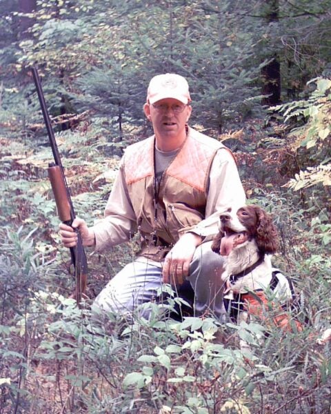 A man in hunting gear kneels in a forest holding a rifle, accompanied by a dog holding a bird in its mouth.