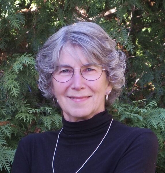 Nancy Jorgensen with glasses, and a black turtleneck stands in front of green foliage, smiling at the camera.
