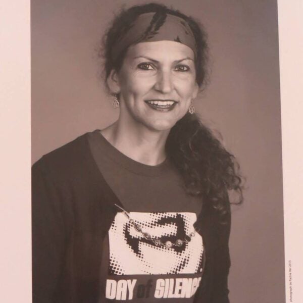 A woman with curly hair wears a headband, earrings, and a Day of Silence T-shirt, smiling at the camera in a black-and-white portrait.