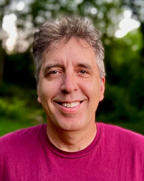 Ken Szymanski with a maroon shirt smiles outdoors with greenery in the blurred background.
