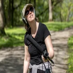 Bridgit Bowden wearing headphones and sunglasses holds a microphone outdoors in a wooded area, looking upward while standing on a sunlit path.