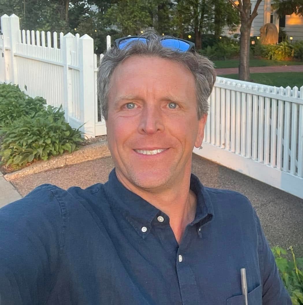 Dan Simmons smiles at the camera outdoors, standing in front of a white picket fence and greenery.