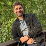 A man with curly hair wearing a dark jacket and watch smiles while leaning on a wooden railing in a wooded outdoor setting.