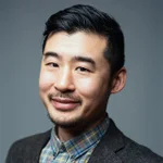 A man with short black hair, a mustache, and a goatee, wearing a plaid shirt and a dark blazer, smiles against a plain gray background.