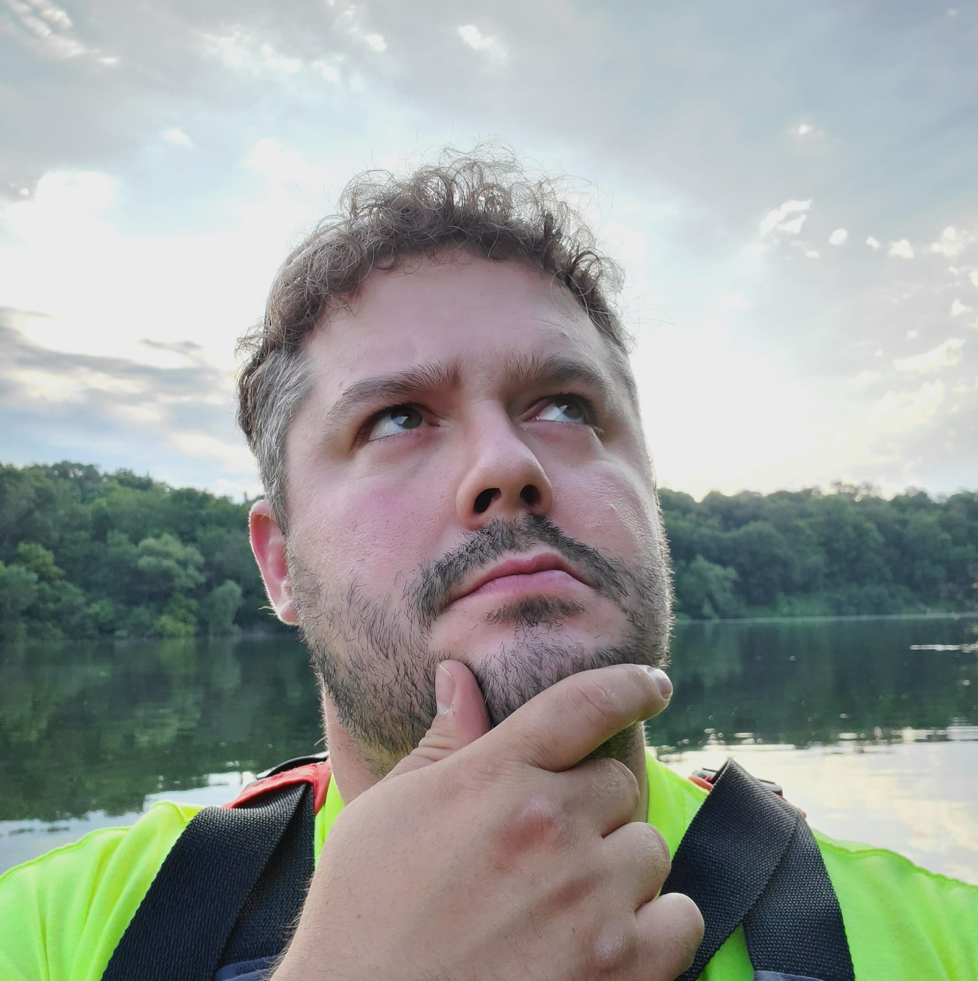 A man with a thoughtful expression stands outdoors near a calm lake, with trees and a cloudy sky in the background.