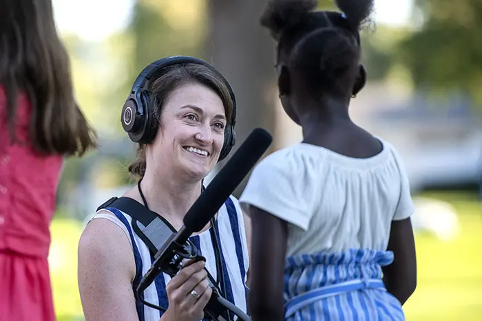 A woman wearing headphones holds a microphone while interviewing a young girl outdoors. Another child stands nearby, partially visible.