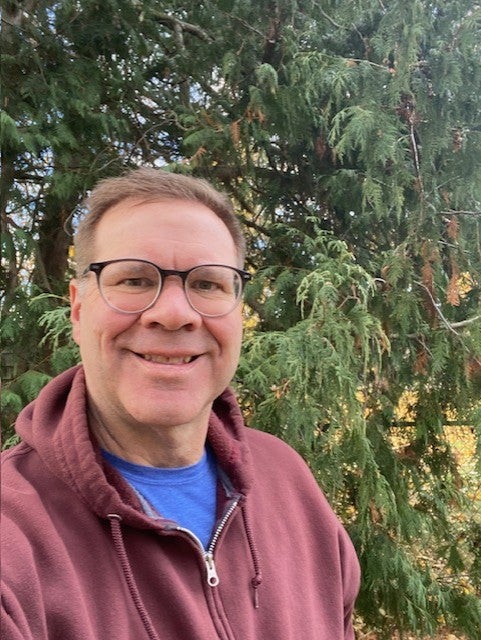 A man wearing glasses and a maroon hoodie smiles in front of green trees outdoors.
