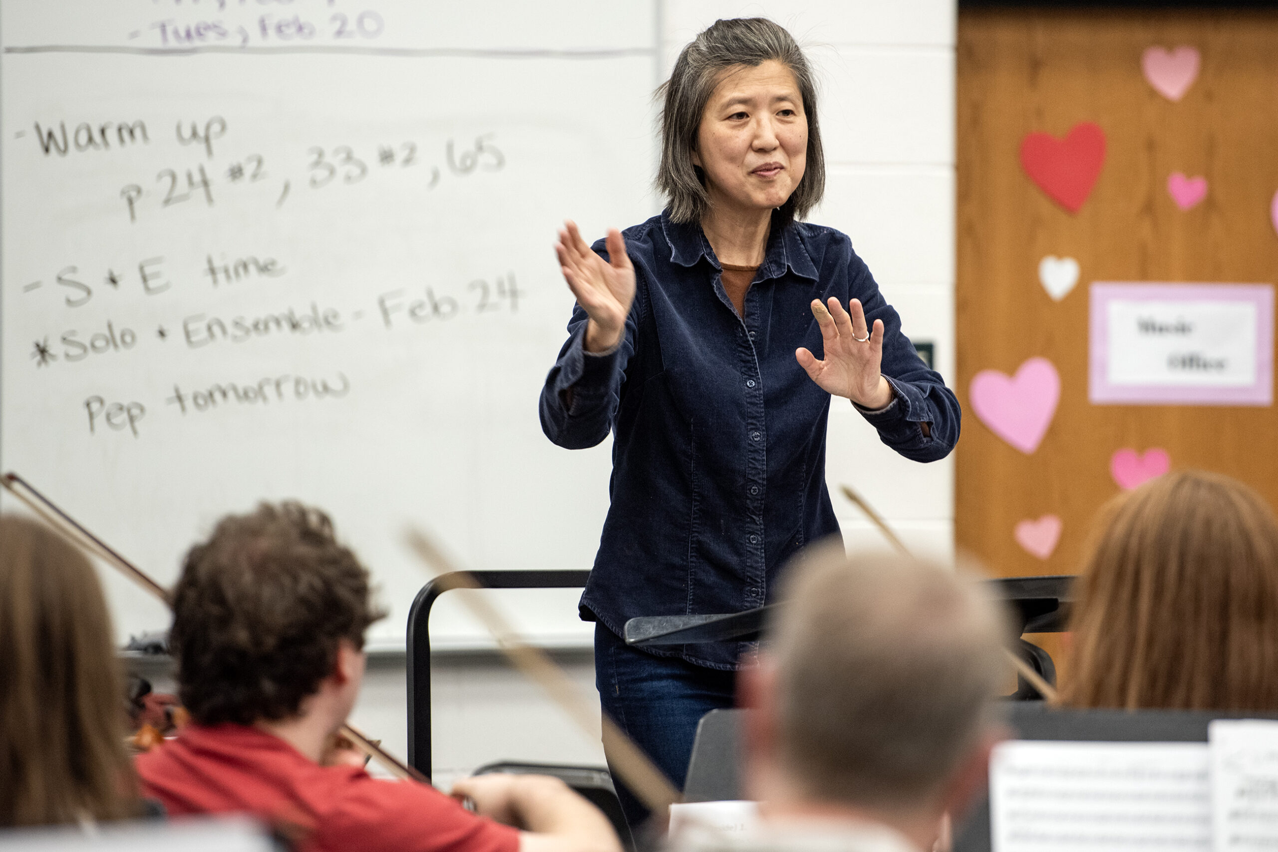 A director smiles and gestures as she stands in front of the group of musicians.