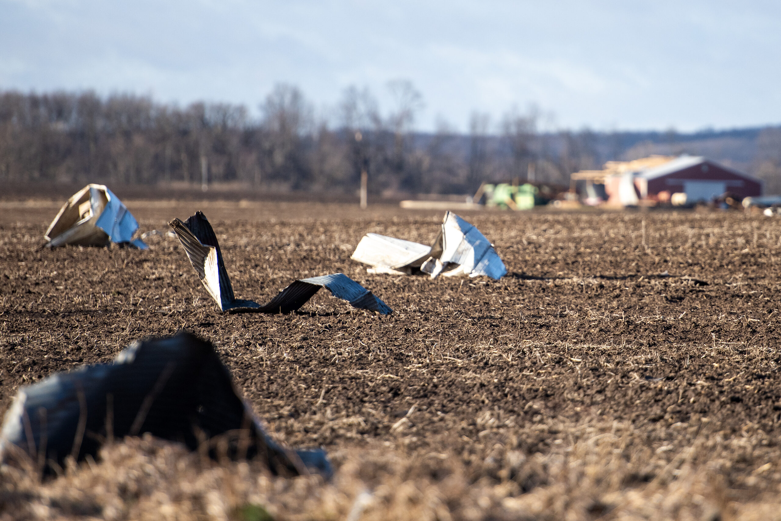 Scraps of structures sit on a large empty field. A barn can be seen in the background.