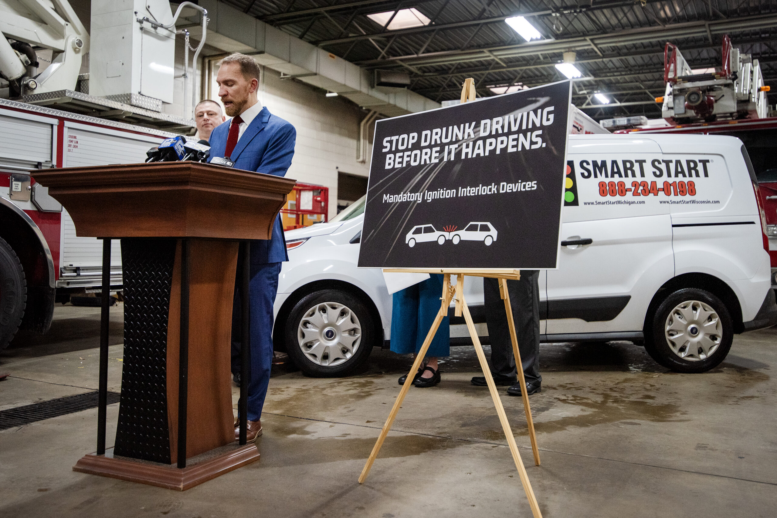 A sign that says "Stop drunk driving before it happens. Mandatory ignition interlock devices." is posted next to Chris Larson as he speaks at a podium.