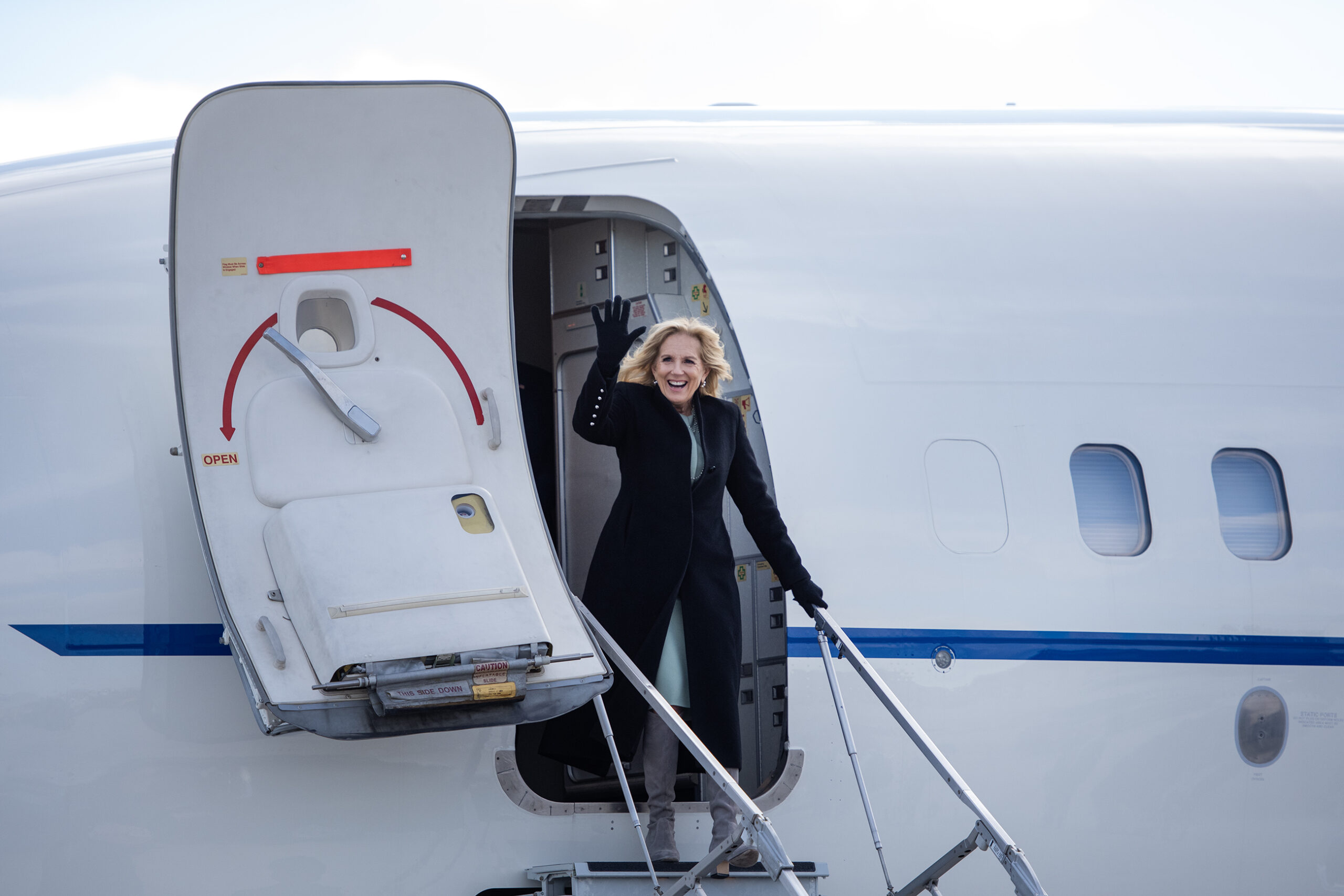 Jill Biden waves as she exits the white aircraft. She wears a black coat and black gloves.