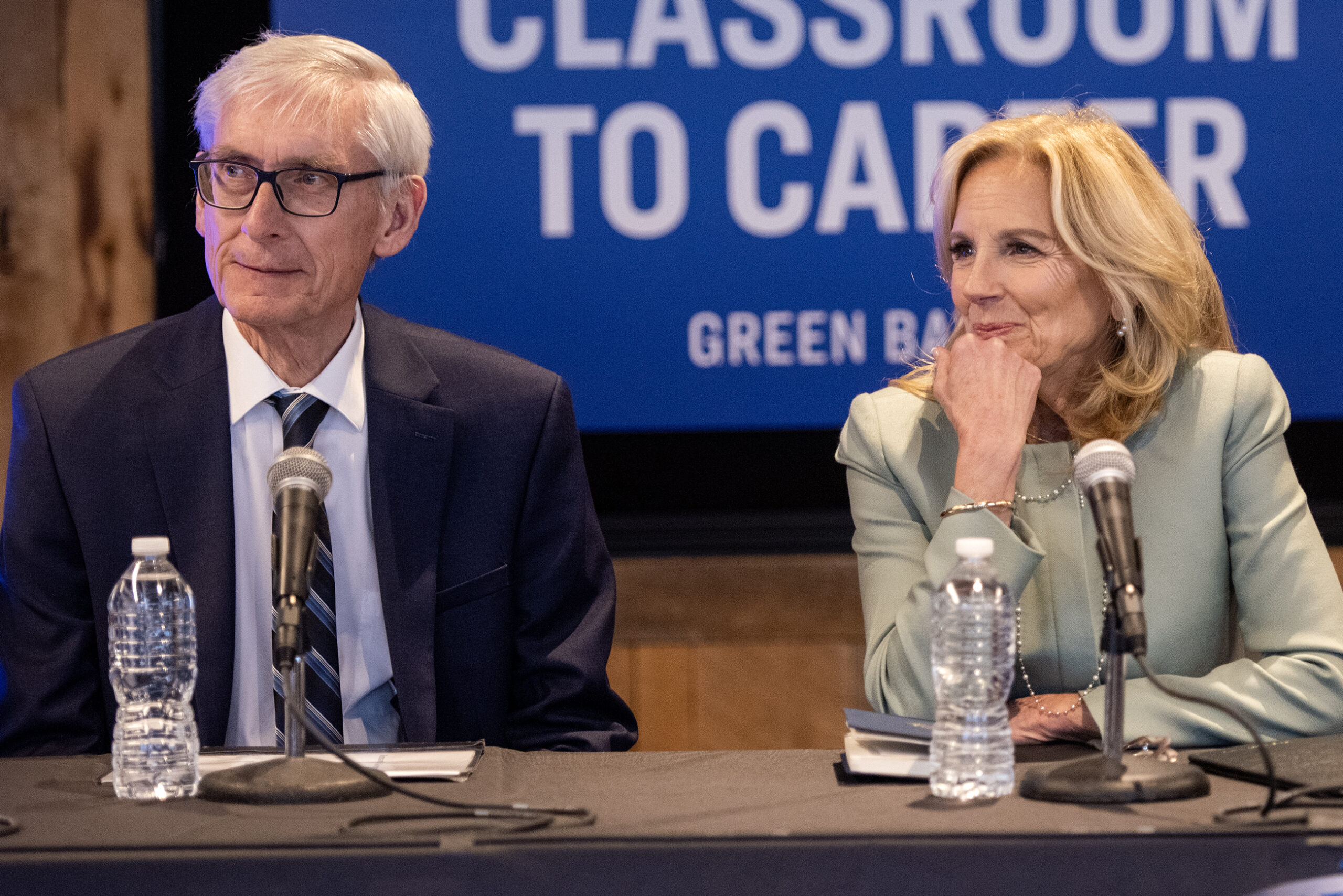 Jill Biden rests her head on her hand as she smiles. Tony Evers sits next to her.
