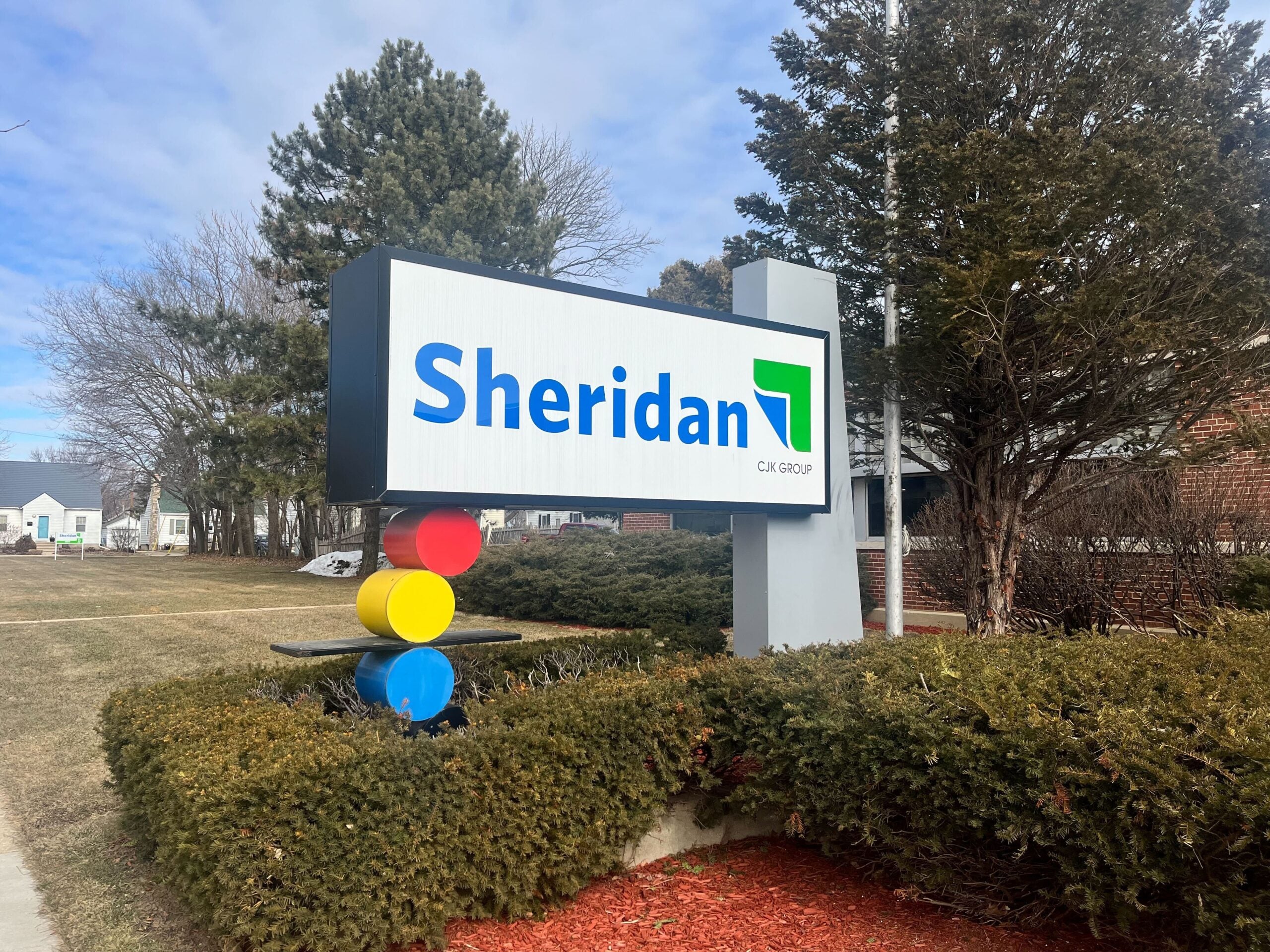 A white sign with blue lettering says "Sheridan." Below the sign are three dots in a vertical line -- red, yellow and blue, respectively. Bushes surround the sign.