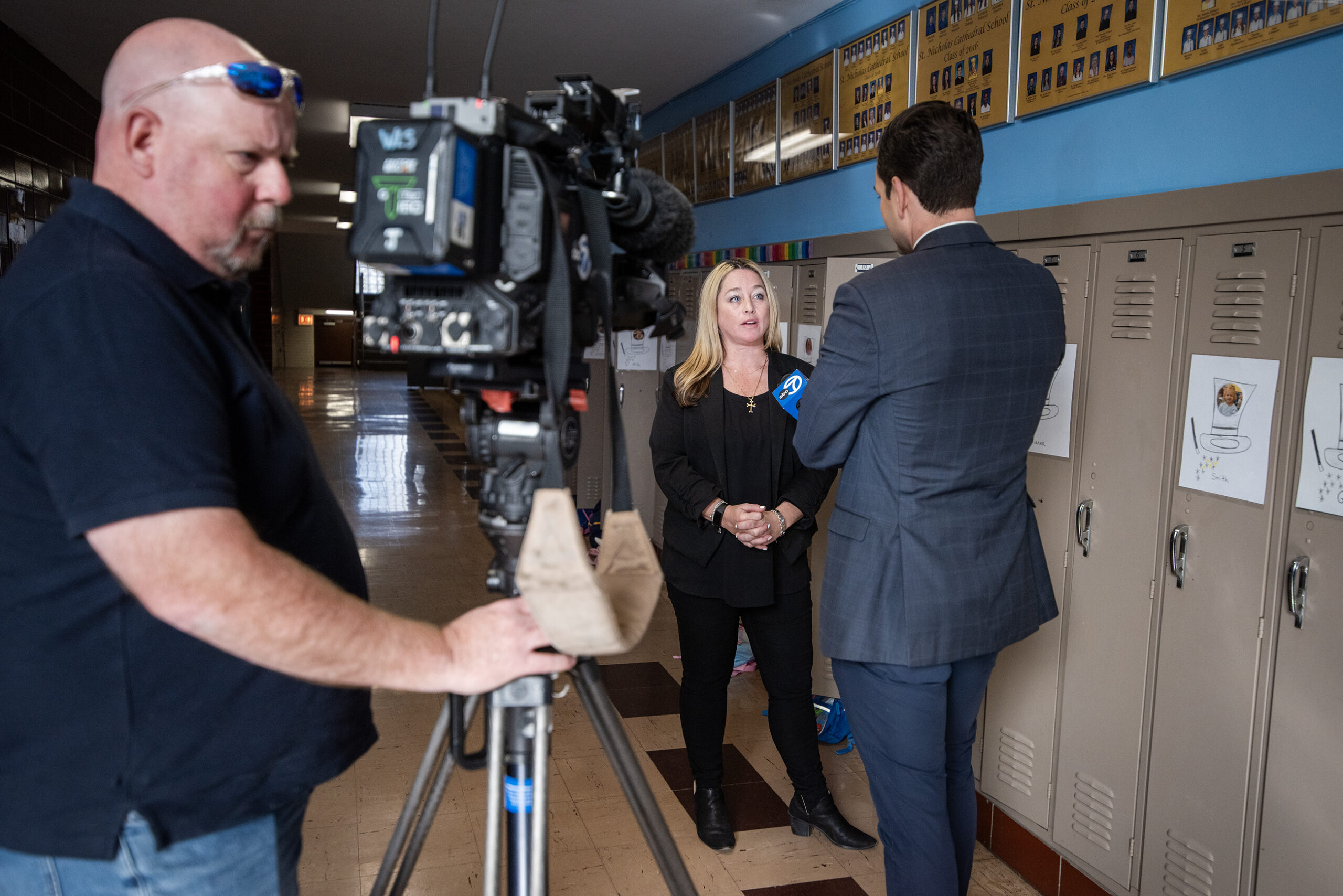 Anna Cirilli speaks with a TV news reporter in front of a camera and lockers inside a school.