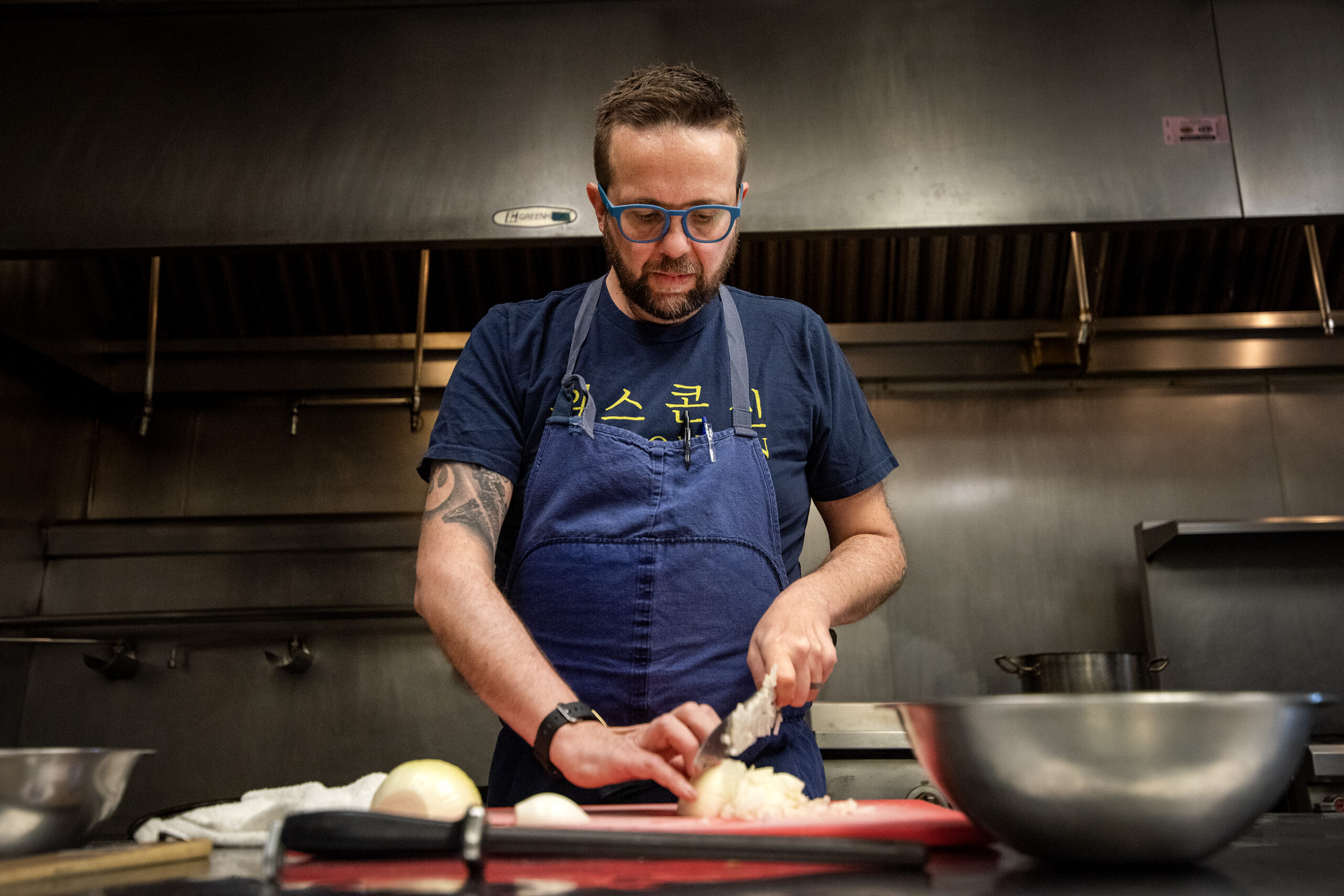 Dan Jacobs wears blue glasses and an apron as he chops an onion.