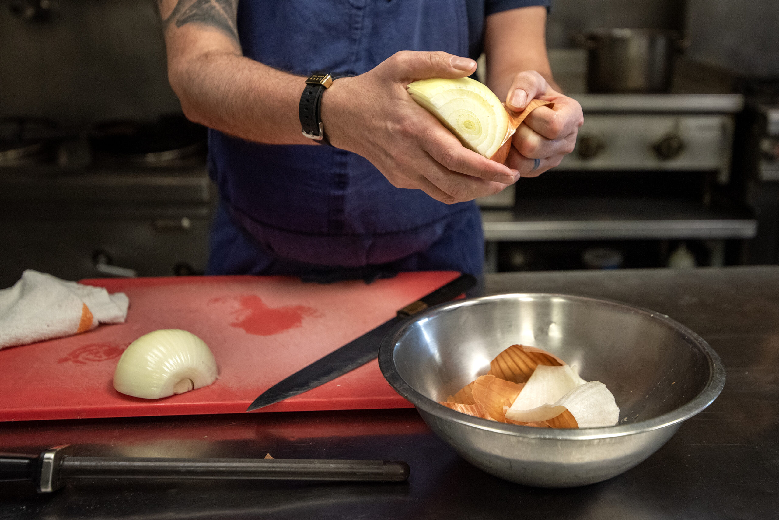 A close up of an onion in Dan Jacobs' hands as he peels it and discards the peel into a bowl.