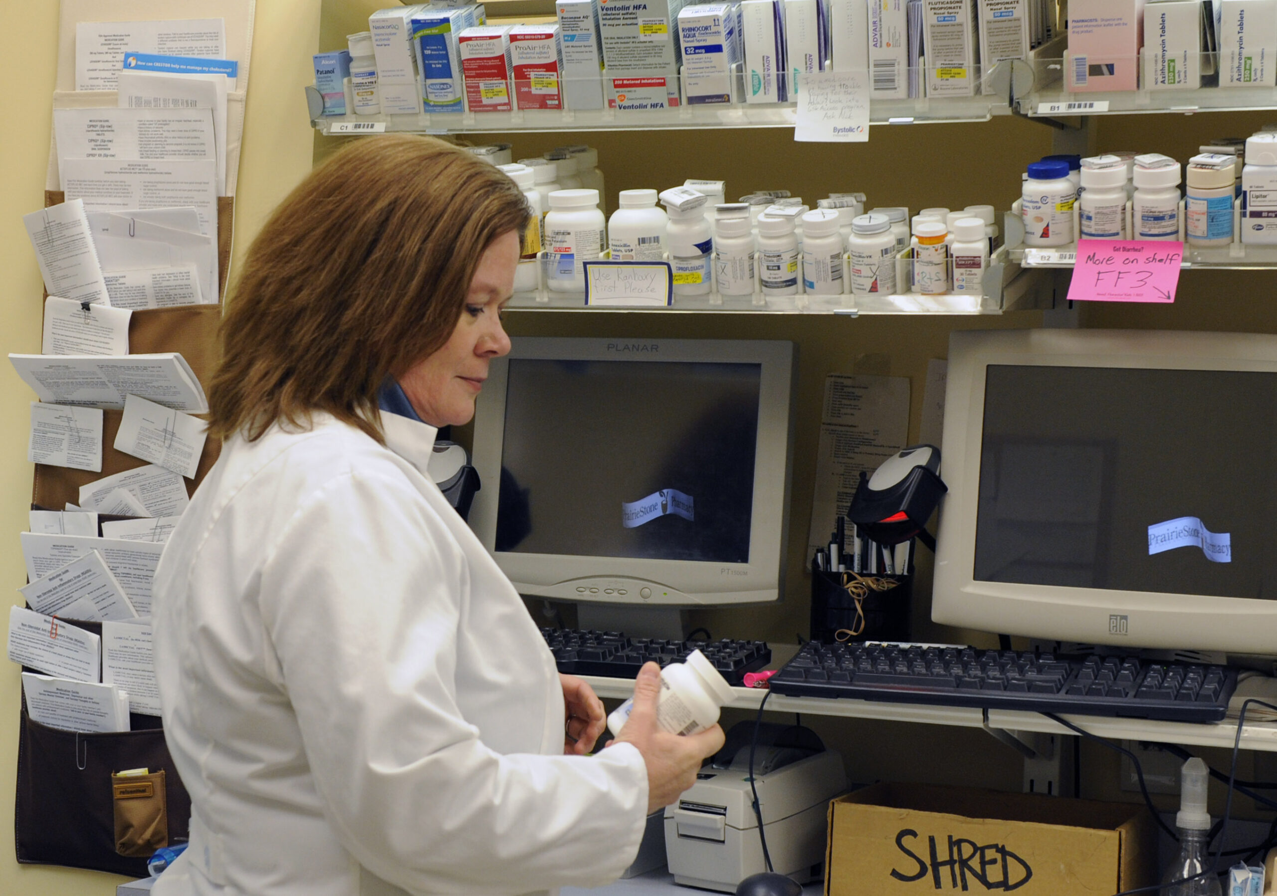 A pharmacist holds a bottle of pills as she stands in front of computers.
