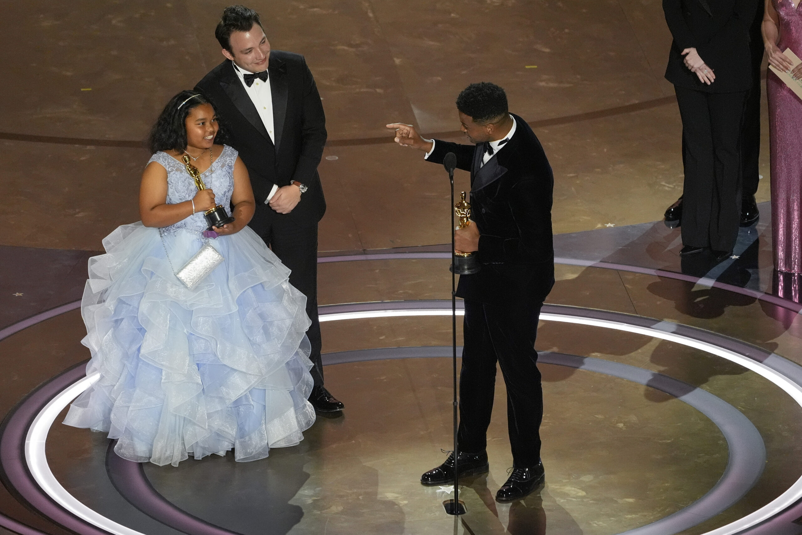 A girl waring a blue dress holds an Oscar on stage after a film she was in won the award. The director of the film is at the microphone and pointing at the girl. 