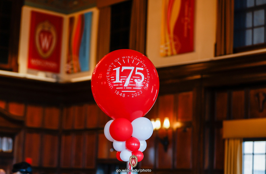 A bright red balloon with 175 printed on it floats above a bouquet of red and white balloons in a dark-paneled room.