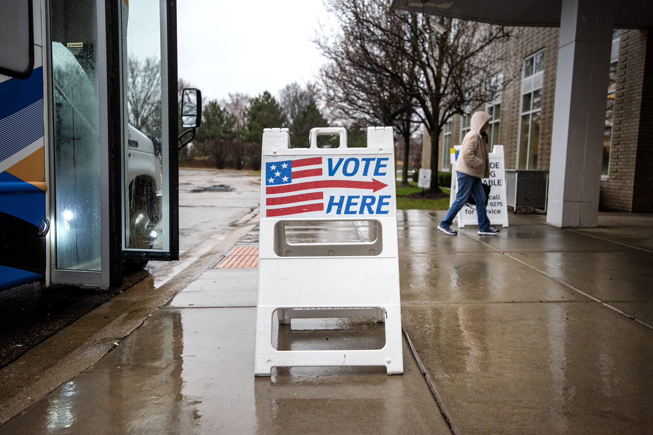 A sign with a US flag that says "vote here" with an arrow is displayed outside on a sidewalk. A parked bus and a pedestrian are nearby.