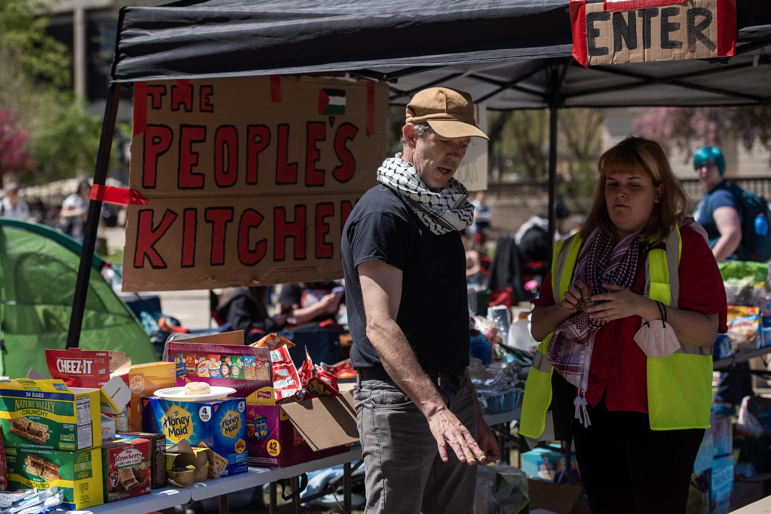 Two people stand under a tent where food is stored. A sign says "the people's kitchen."