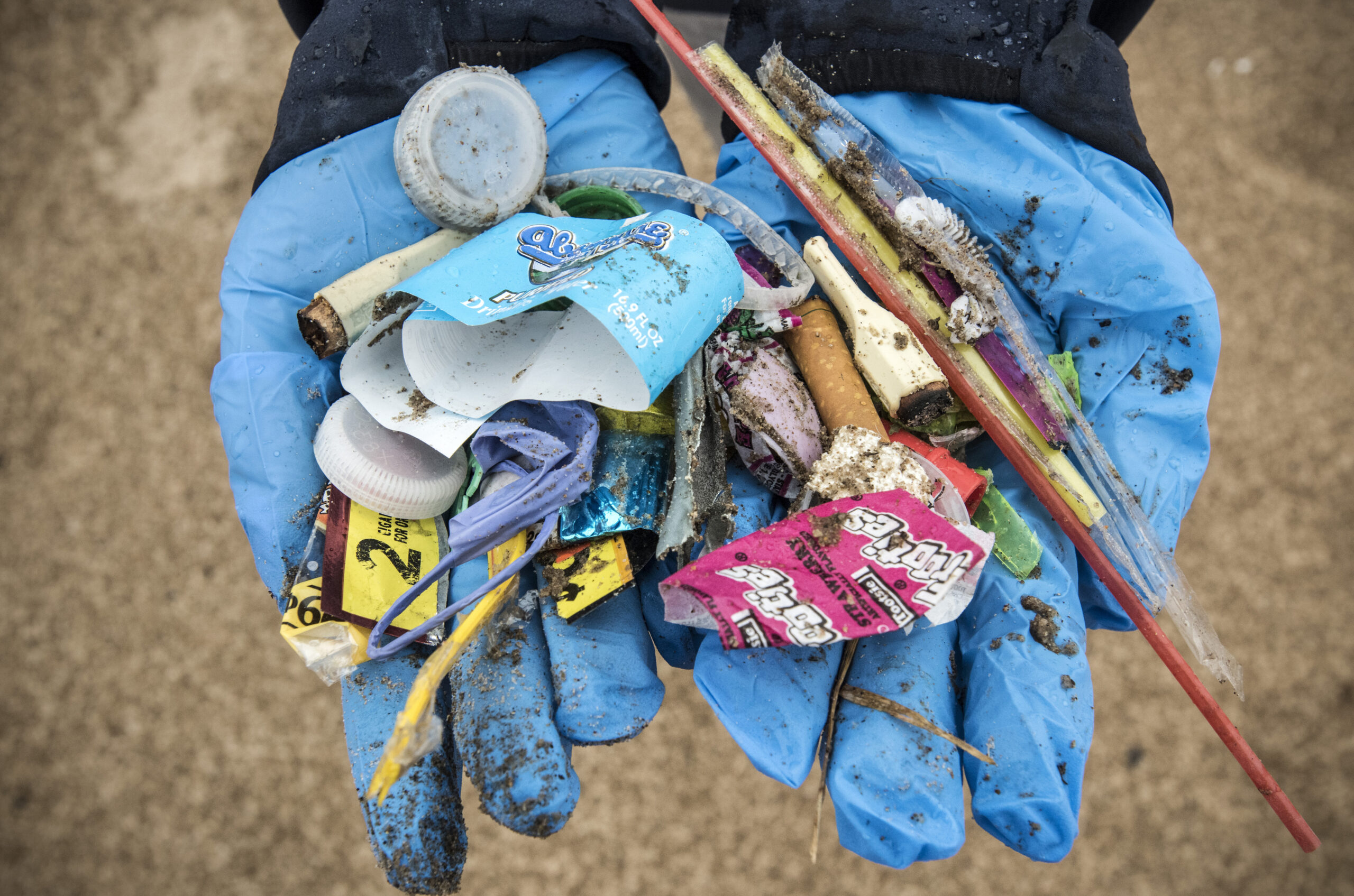 Gloved hands hold bottle caps, food wrappers and other trash found on the beach