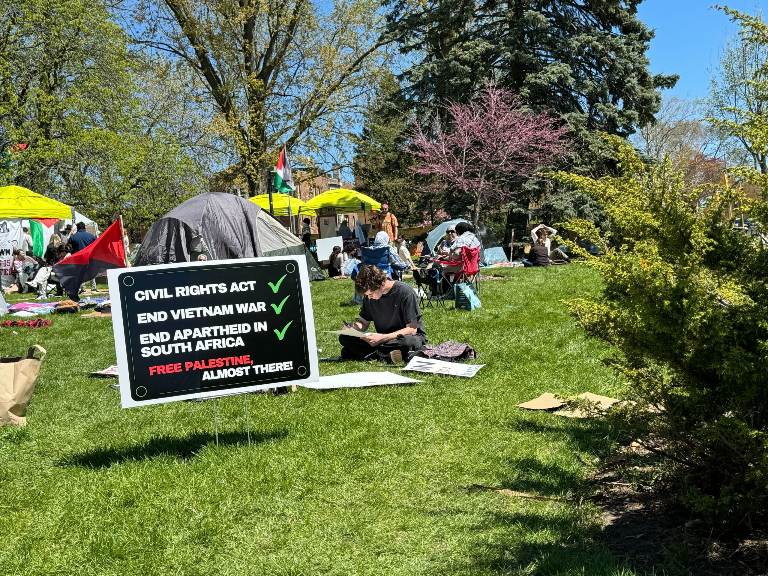 A black sign in the grass reads "Civil Rights Act (checkmark, End Vietnam War (checkmark), End apartheid in South Africa (checkmark), Free Palestine, almost there!" Behind the sign are tents and lawn chairs with people scattered about.
