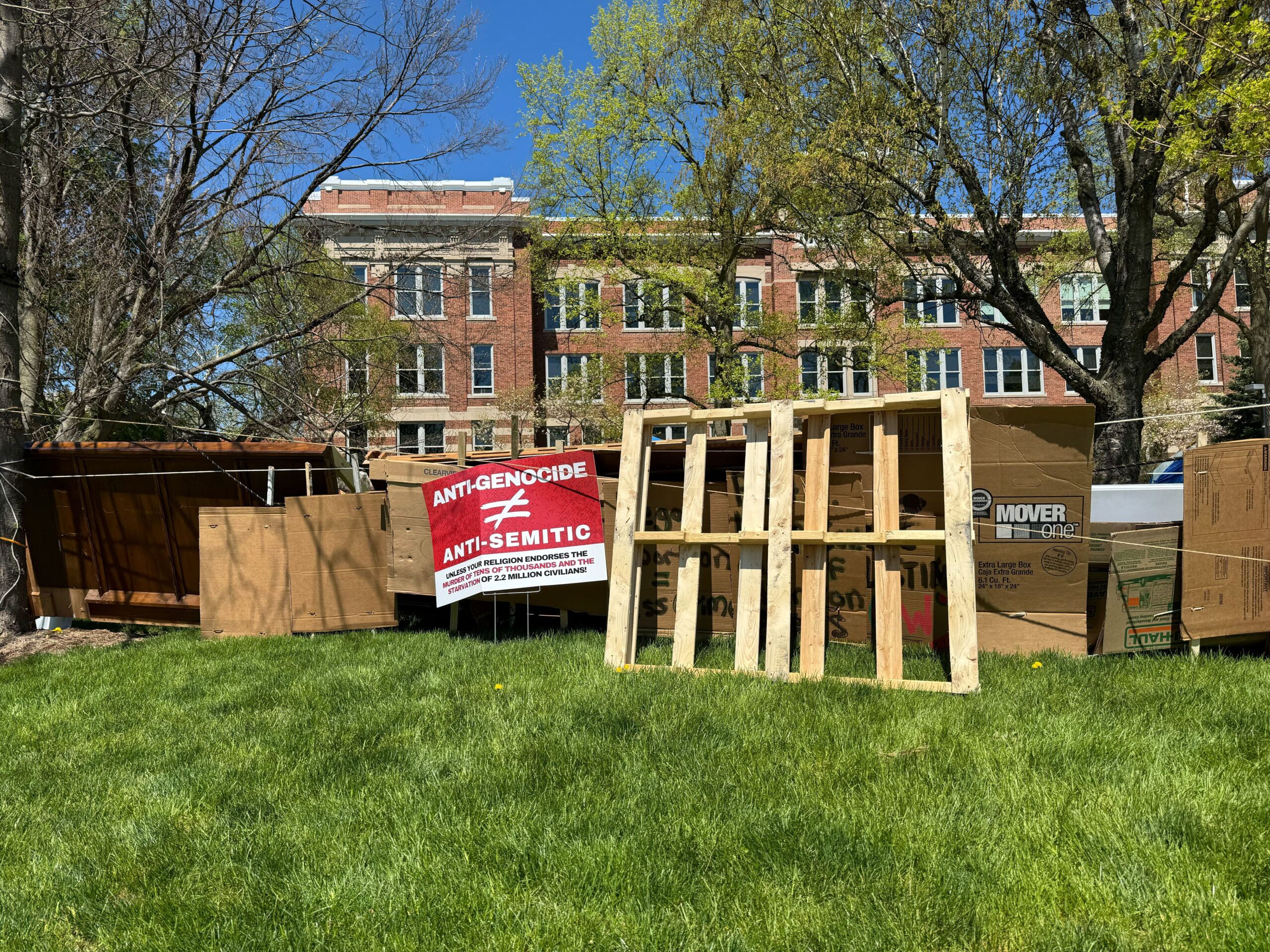 Plywood, folding tables and cardboard create a barrier in the grass on UW-Milwaukee's campus. A brick campus building is in the background with a blue sky.