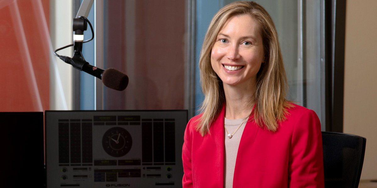 A photograph of a smiling woman in a bright red jacket sitting in Wisconsin Public Radio's studios in Madison. The woman is WPR Director Sarah Ashworth.