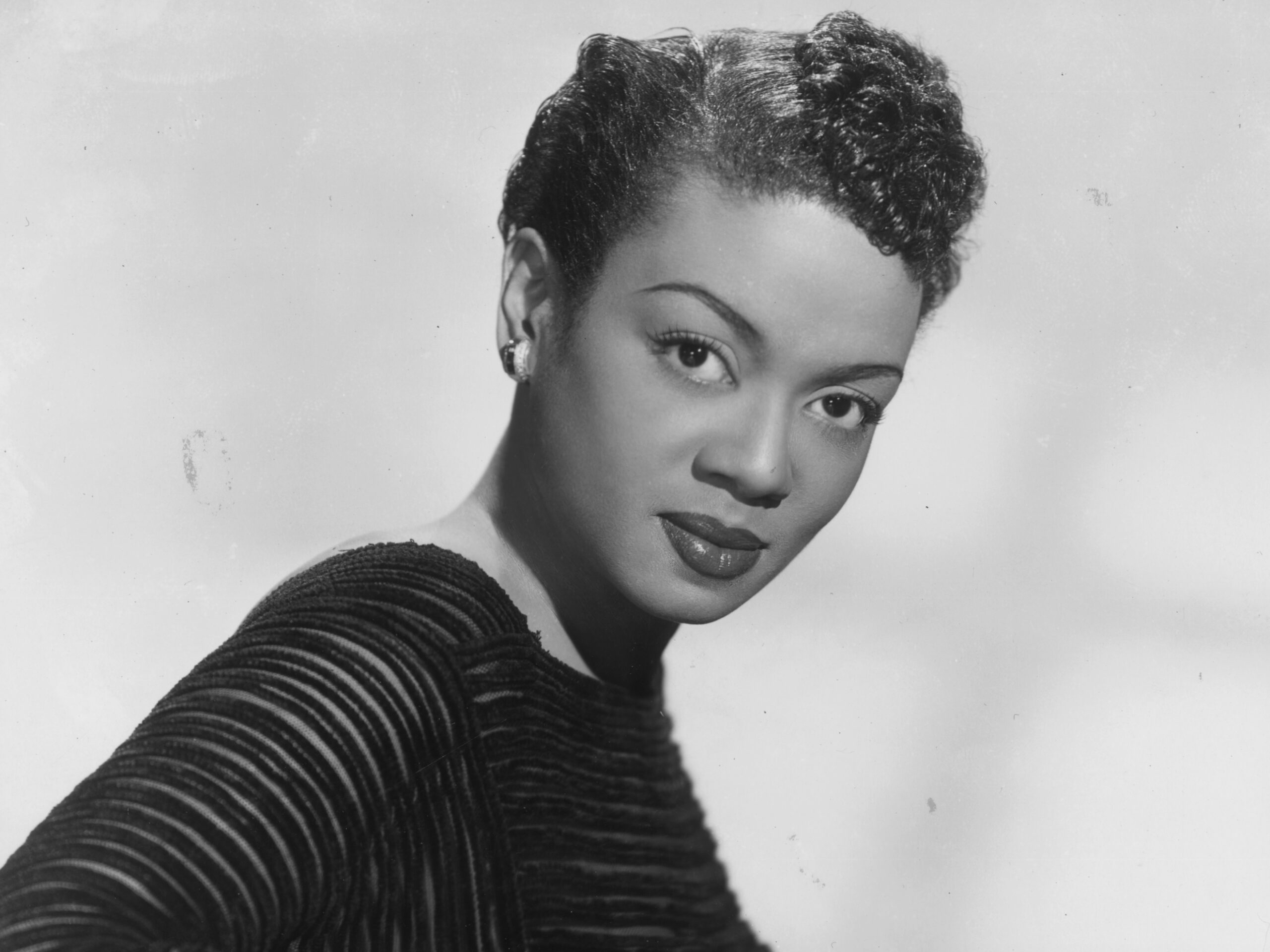 circa 1955: Studio portrait of Trinidadian-born jazz pianist and vocalist Hazel Scott, wearing a black dress with thin sheer panels, leaning against a white piano. Scott trained at the Julliard School. (Photo by Hulton Archive/Getty Images)
