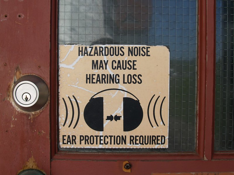 A sign reads "Hazardous Noise May Cause Hearing Loss, Ear Protection Required." The sign is on a door.