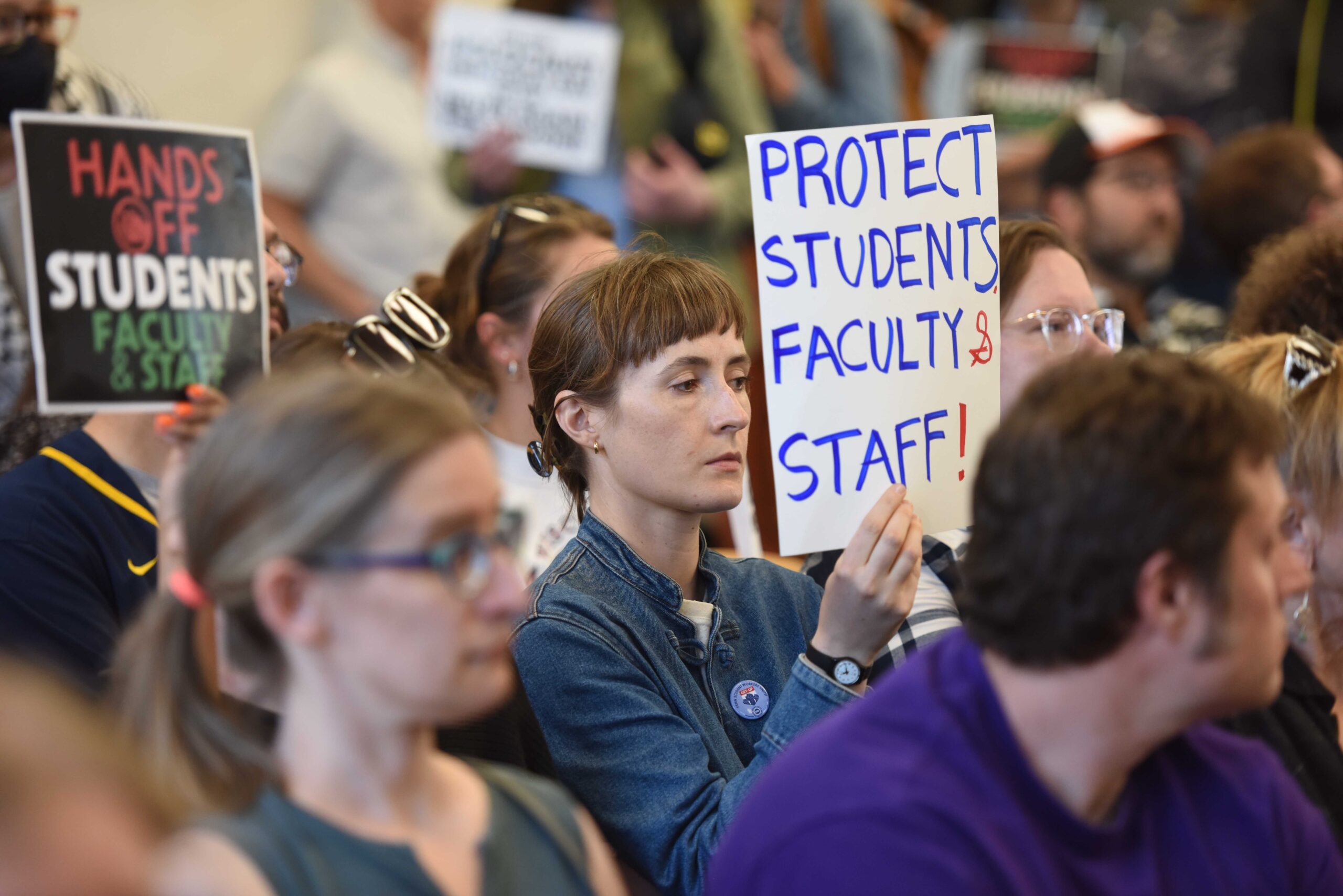 A person in a denim shirt holds a white sign that says "Protects students faculty & staff!" in blue letters.