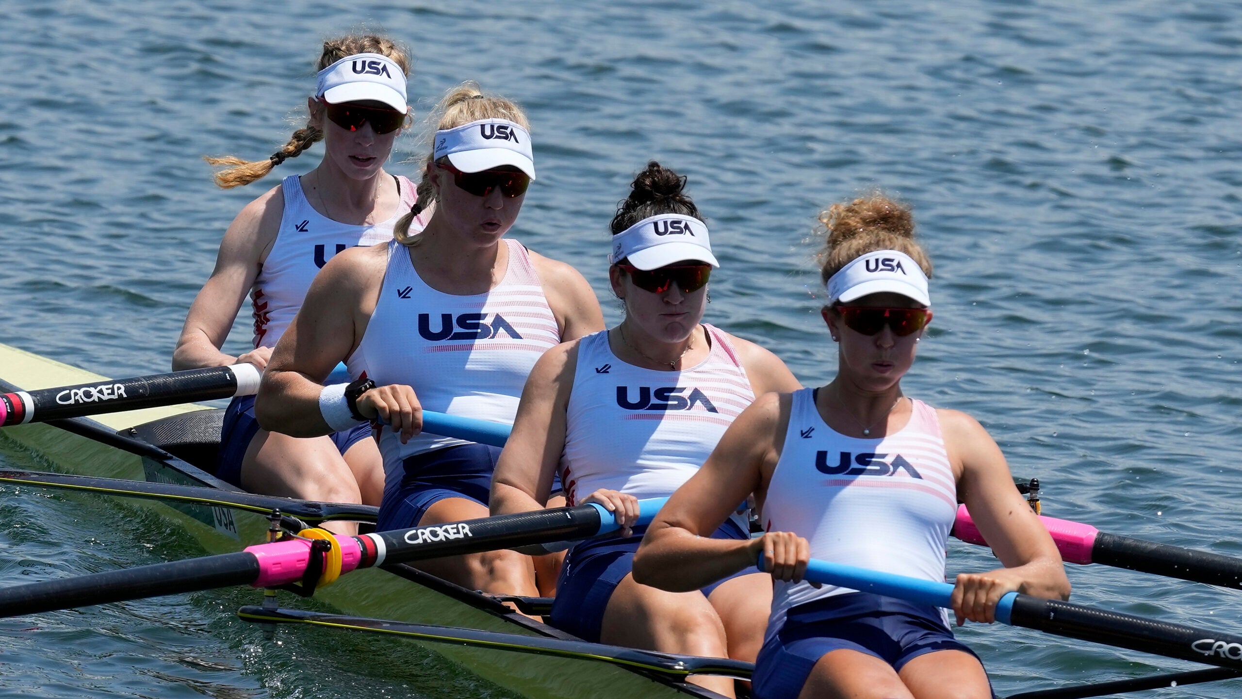 For women row together in a slim rowing boat. They're wearing matching uniforms that read "USA."