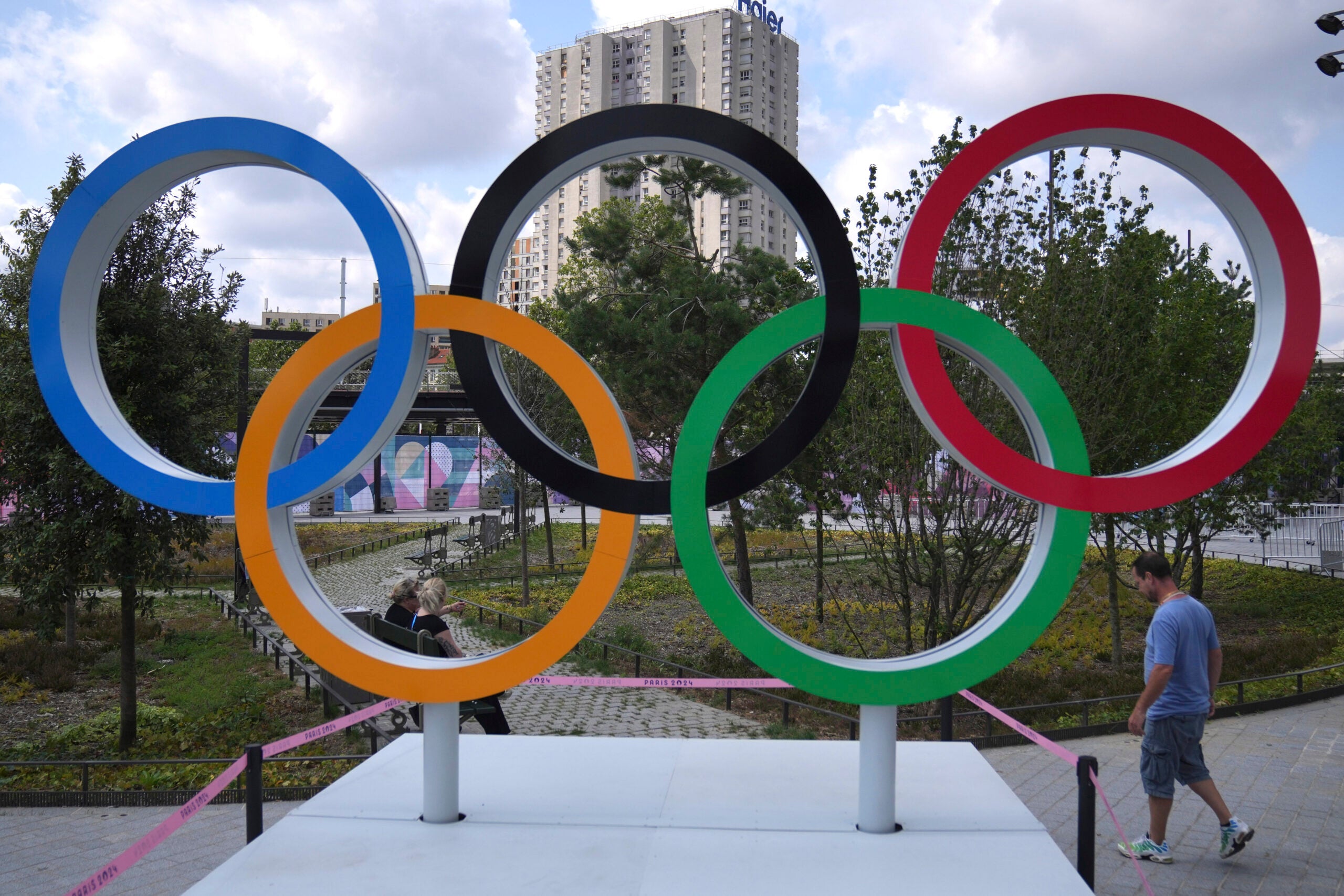 The olympic rings in front of a tall concrete building and some trees.