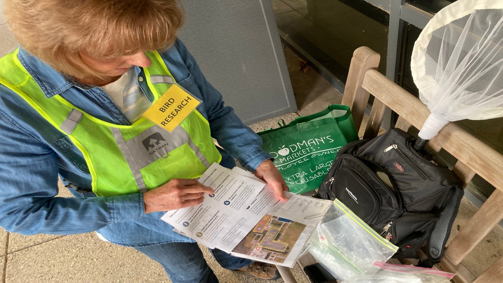 Volunteer Ann Mader wears a bright vest and gathers materials to survey birds. She holds a map of the site, and has a backpack, canvas bag and net beside her.