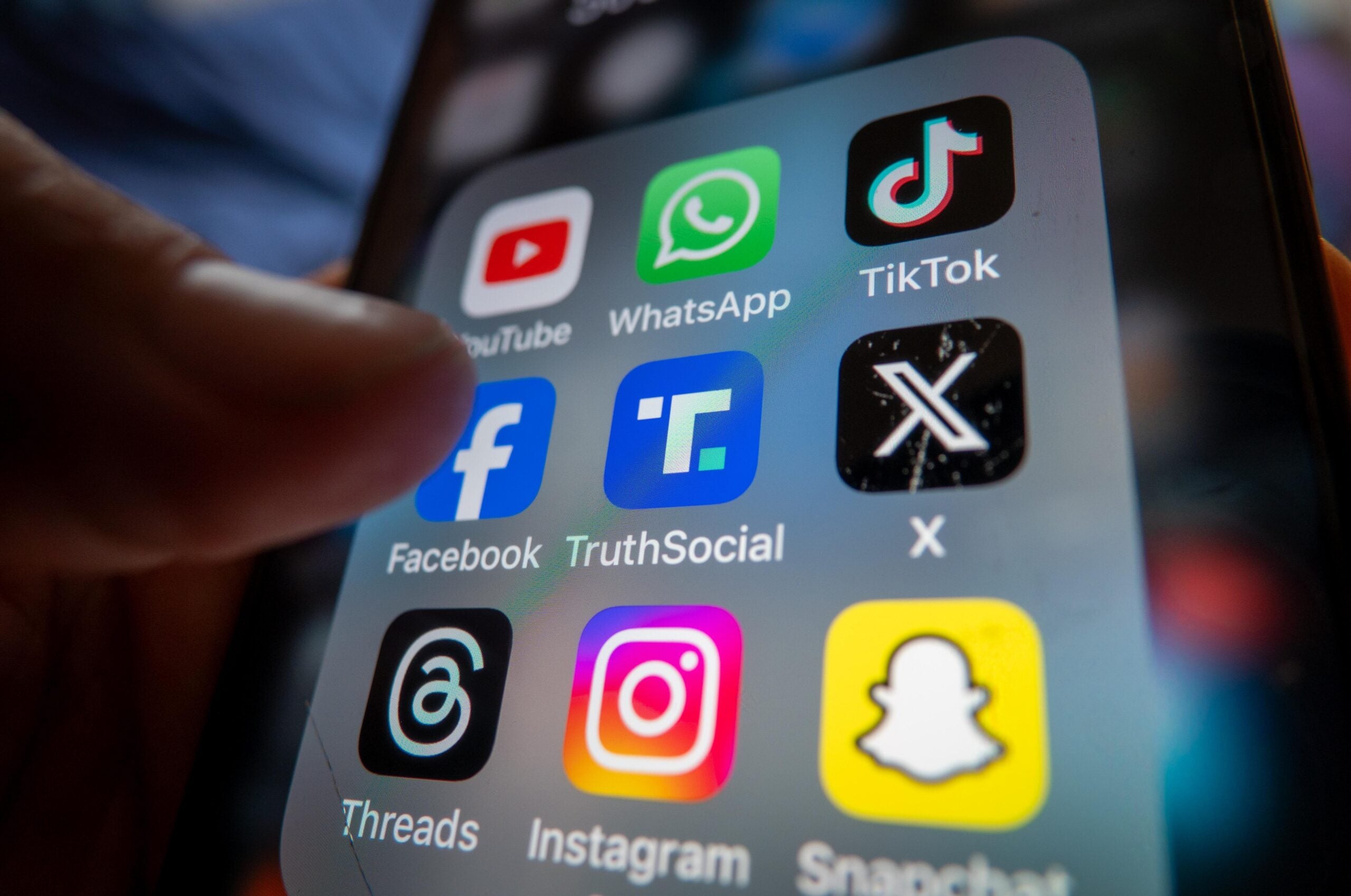 A close-up of a smartphone screen showing various social media app icons, including YouTube, WhatsApp, TikTok, Facebook, Truth Social, X, Threads, Instagram, and Snapchat.