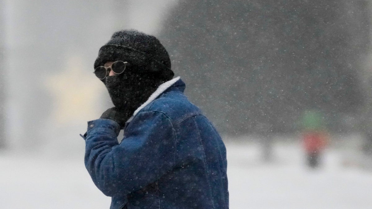 A man walks across the street with a hat, scarf and sunglasses on.
