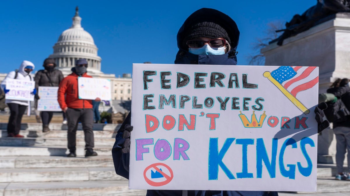 A person holds a sign reading Federal Employees Dont Work for Kings in front of the U.S. Capitol, with others holding signs in the background.