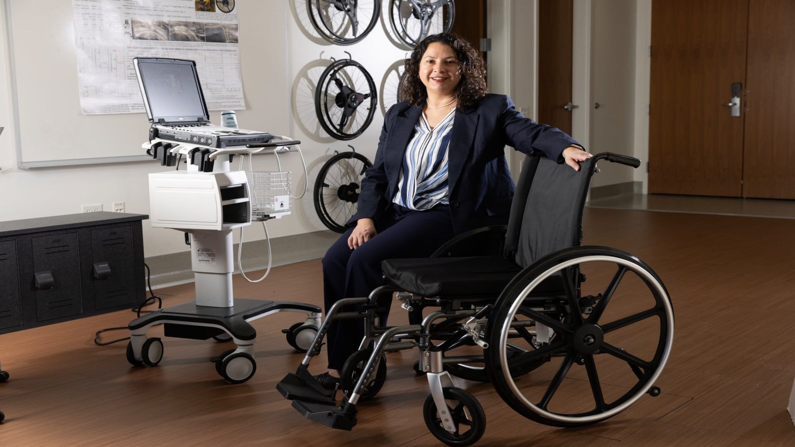 A woman in a blue suit sits on the armrest of a wheelchair, next to a medical device with a monitor, in a professional setting.