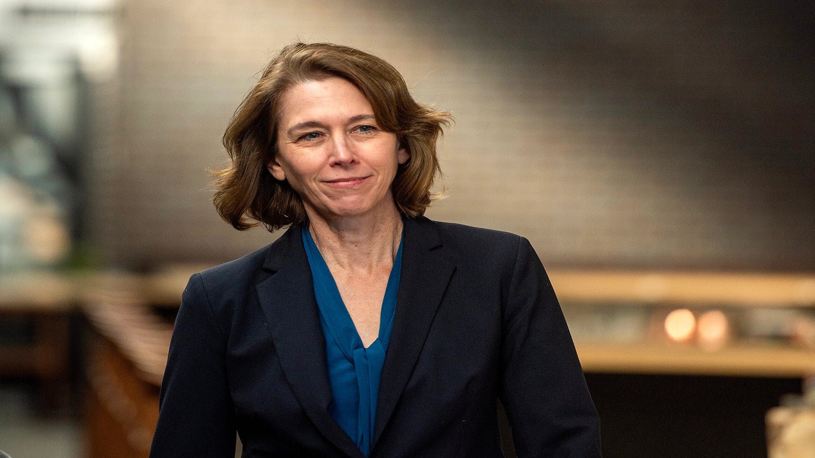 Woman with shoulder-length brown hair wearing a navy blazer and blue blouse, standing indoors with a blurred brick wall background.
