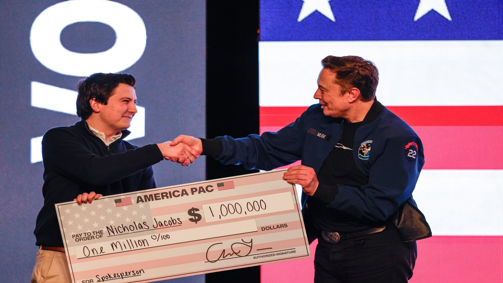 Two men shaking hands on stage, one holding a large novelty check for one million dollars with Nicholas Jacobs written on it, against a backdrop of the American flag.