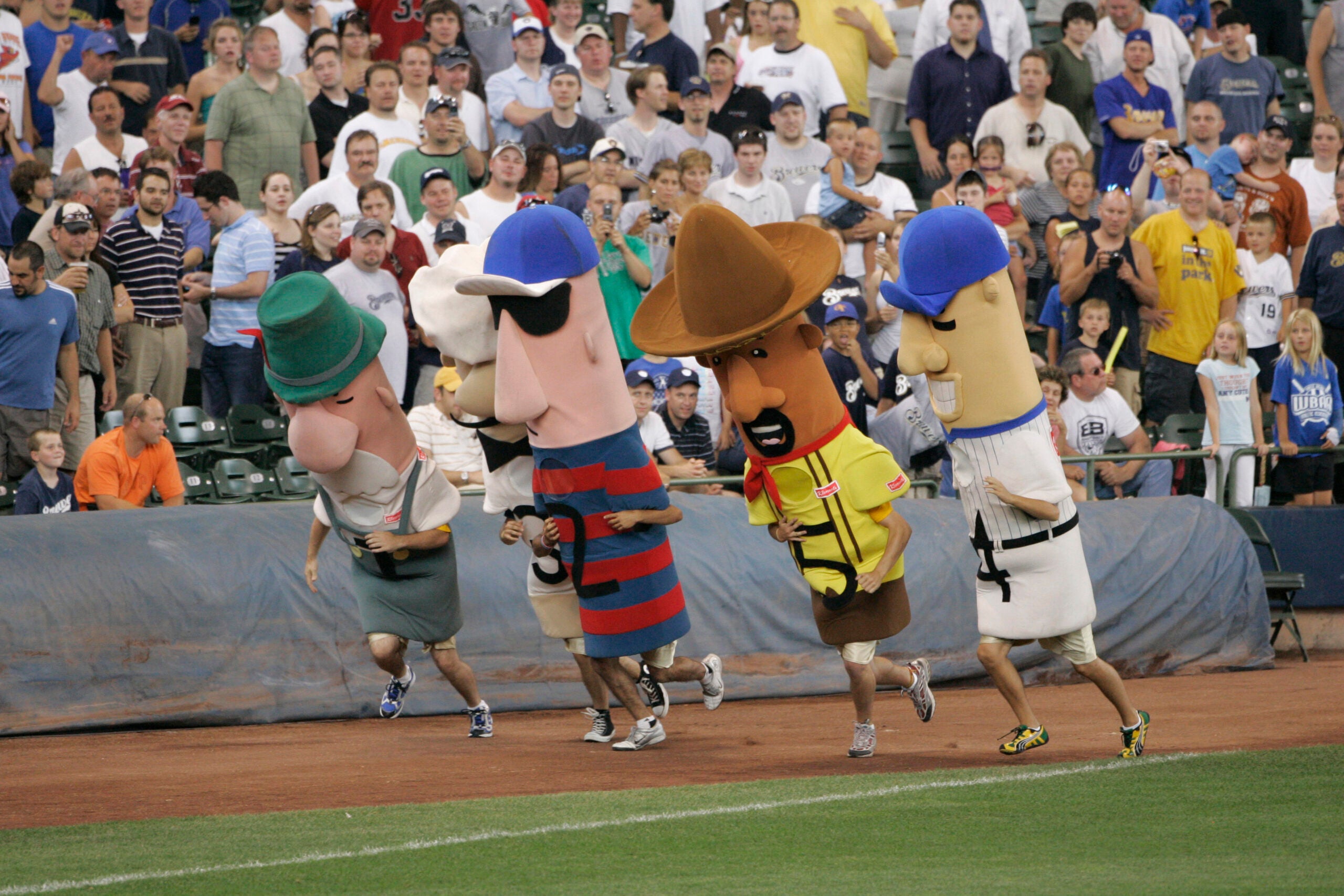 A group of people in large sausage costumes race on a baseball field, with a crowd watching from the stands in the background.