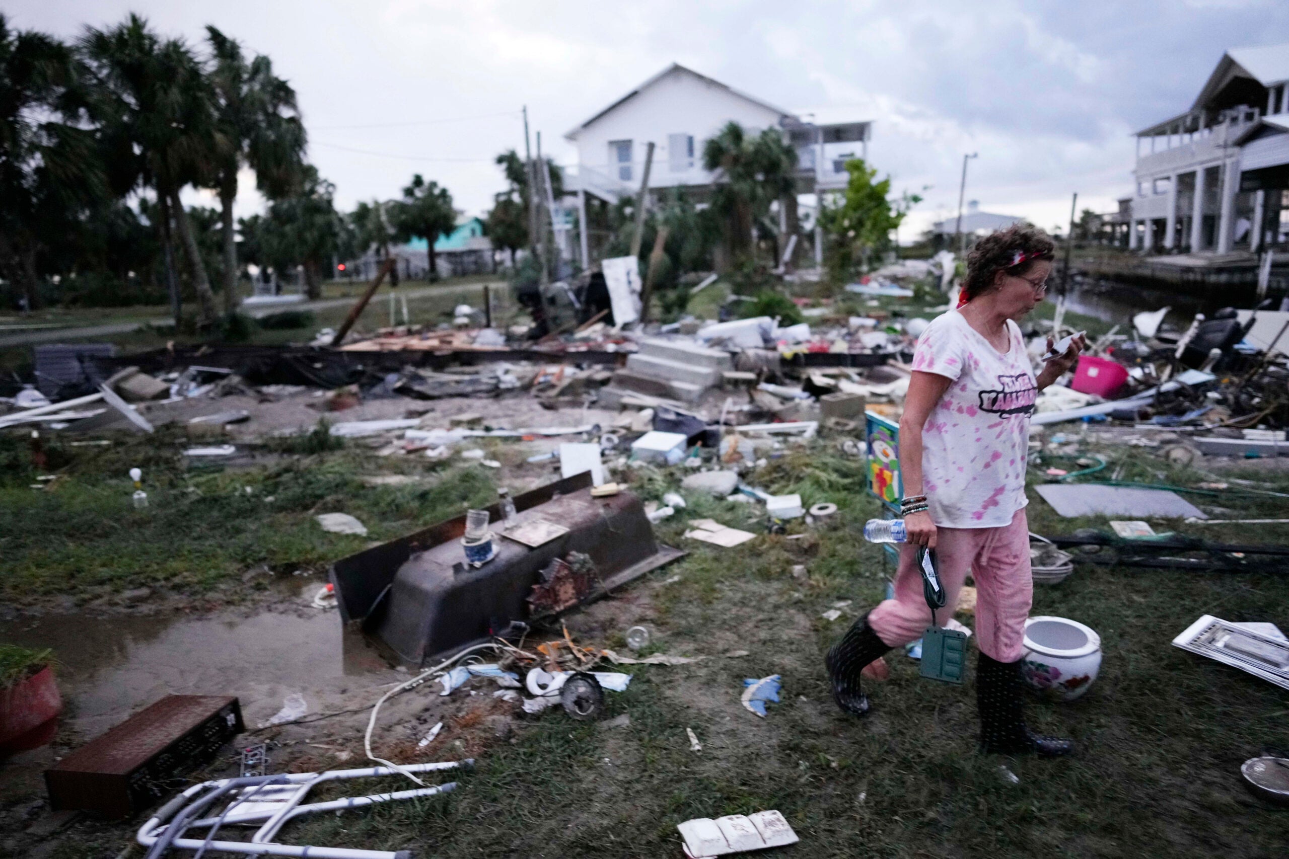A person walks among debris and destruction left by a storm, with damaged houses and scattered belongings in the background.