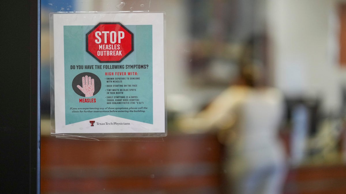 A sign on a glass door warns of a measles outbreak, listing symptoms like fever and rash. A person is blurred in the background, standing near a counter.