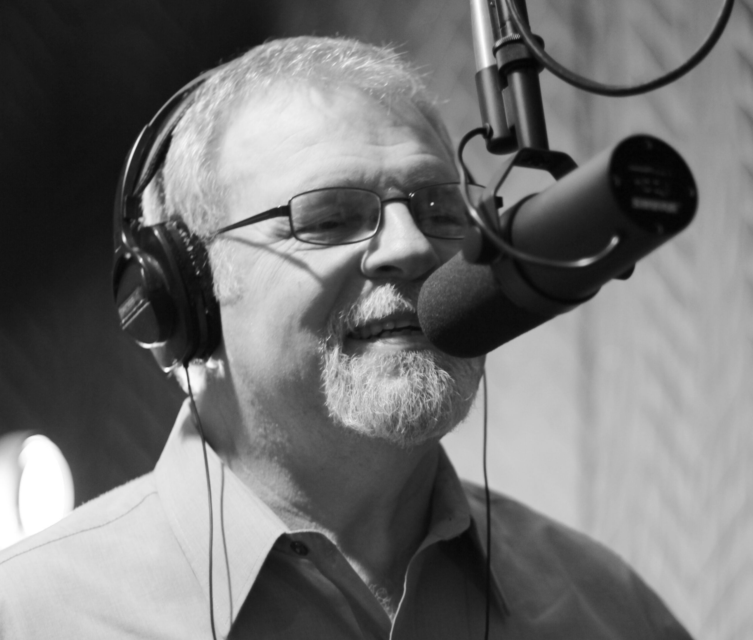 A man wearing headphones and glasses speaks into a microphone in a recording studio.