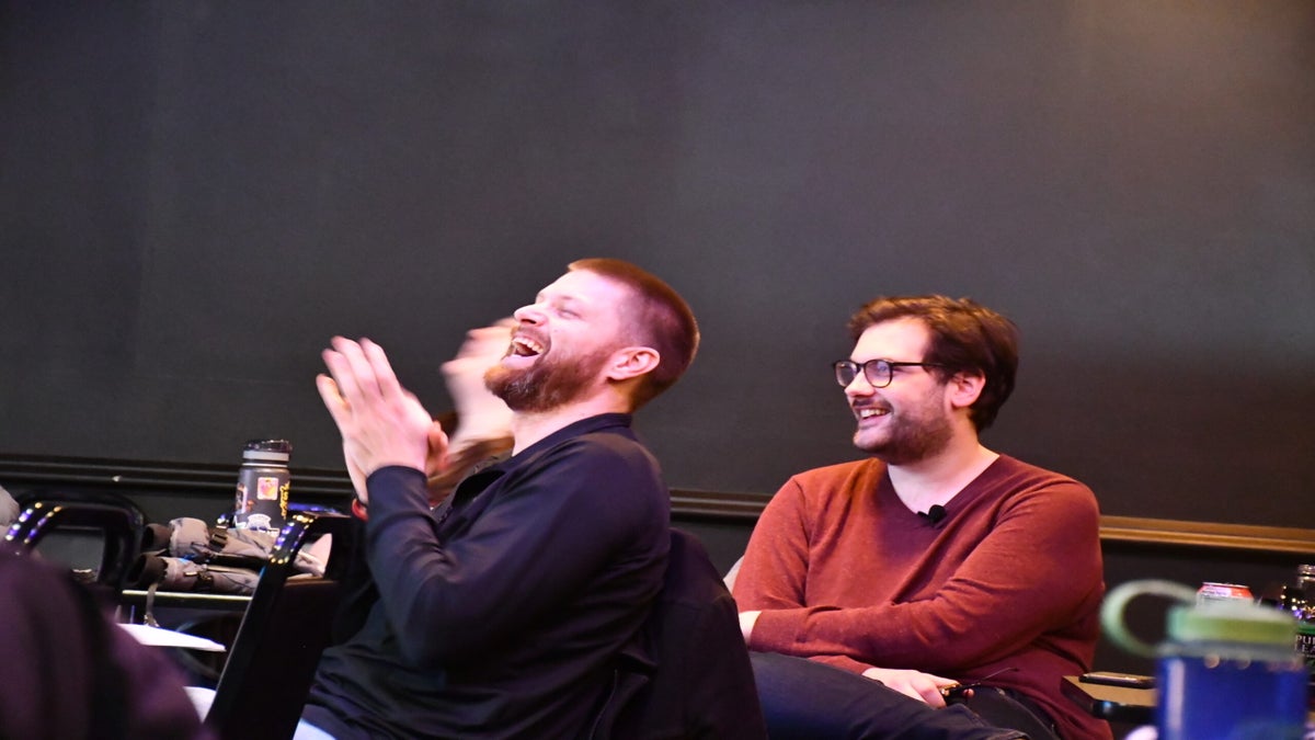 Two men sitting in chairs, both laughing. The man on the left claps his hands while seated. The man on the right wears glasses. Both appear amused.