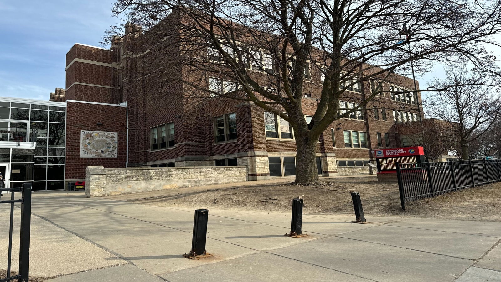 A large, multi-story brick building stands with a tree in front, surrounded by a metal fence and a paved walkway.