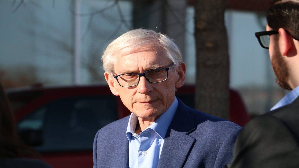 An older man with white hair and glasses wearing a blue suit jacket stands outdoors, facing another person.