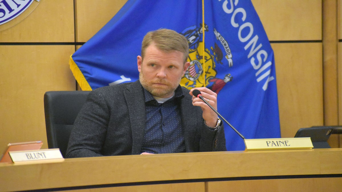 A person seated at a desk with a nameplate Paine, holding a microphone, with a Wisconsin flag in the background.