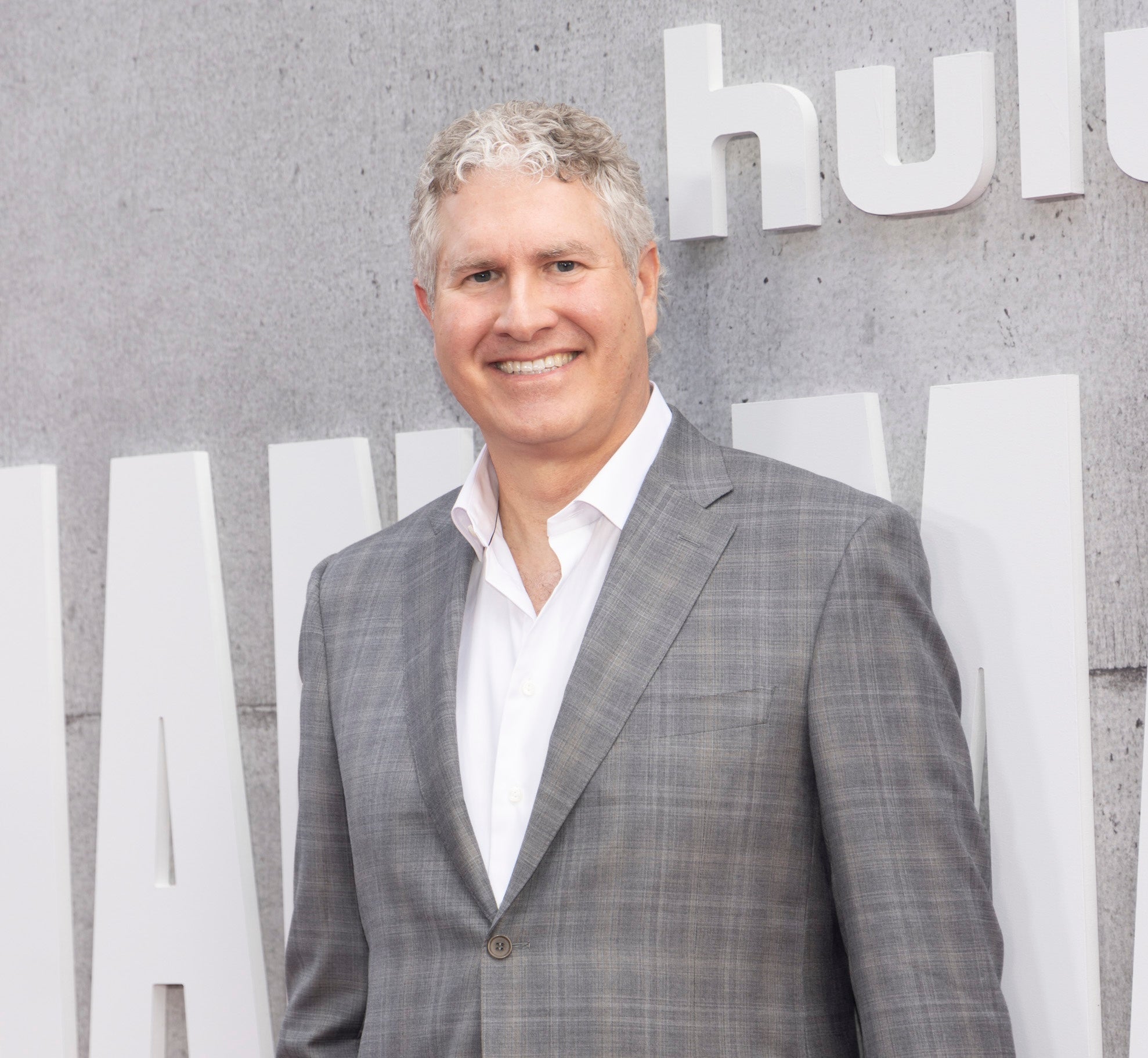 A man in a gray plaid suit and white shirt smiles at a Hulu event, standing in front of a textured gray wall with large signage.